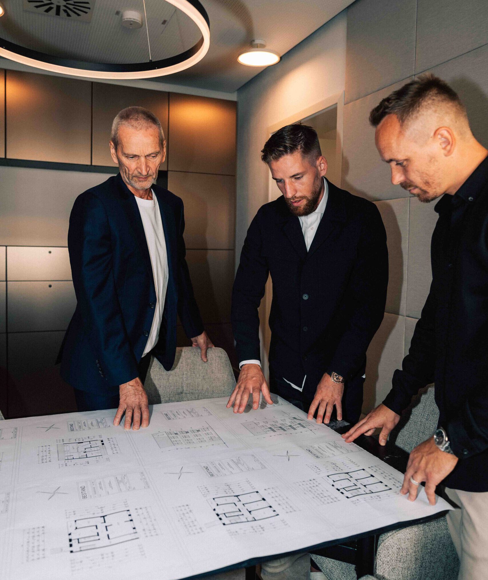Foto von Günter Pusch, Kolja Pusch und Arne Feick, die Teil der Pusch Immobiliengruppe sind. Sie stehen nebeneinander in einem Büro und schauen sich einen Bauplan sowie Grundrisse an.